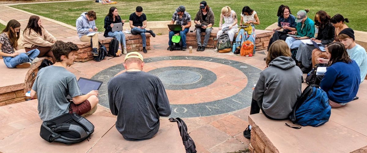 A philosophy class meets outdoors, seated in a circle around a stone plaza on Colorado State University's campus, with students reading, writing, and using laptops during discussion.