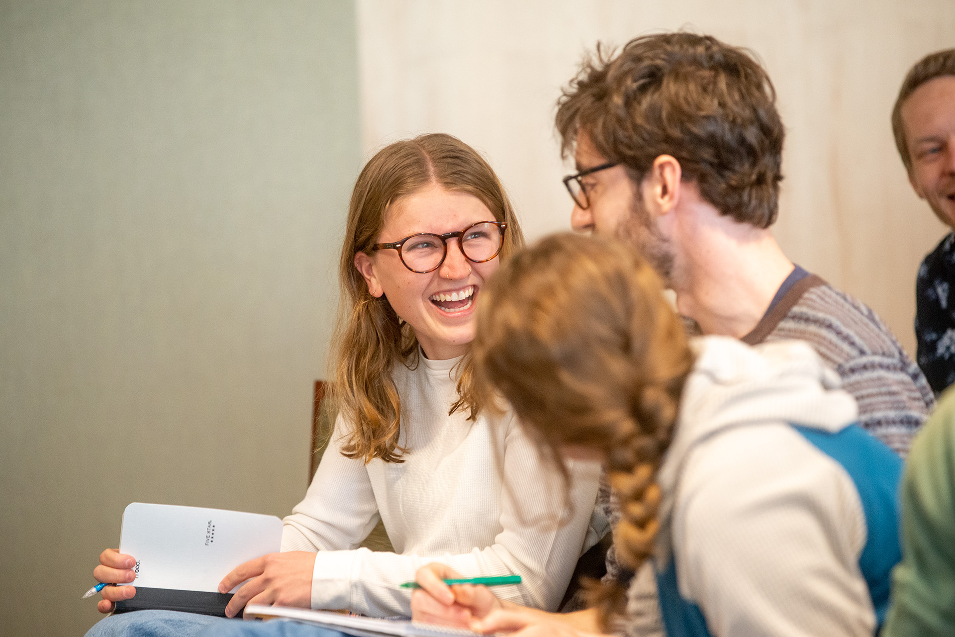 A student laughs warmly during a philosophy class discussion, notebook in hand, surrounded by classmates.