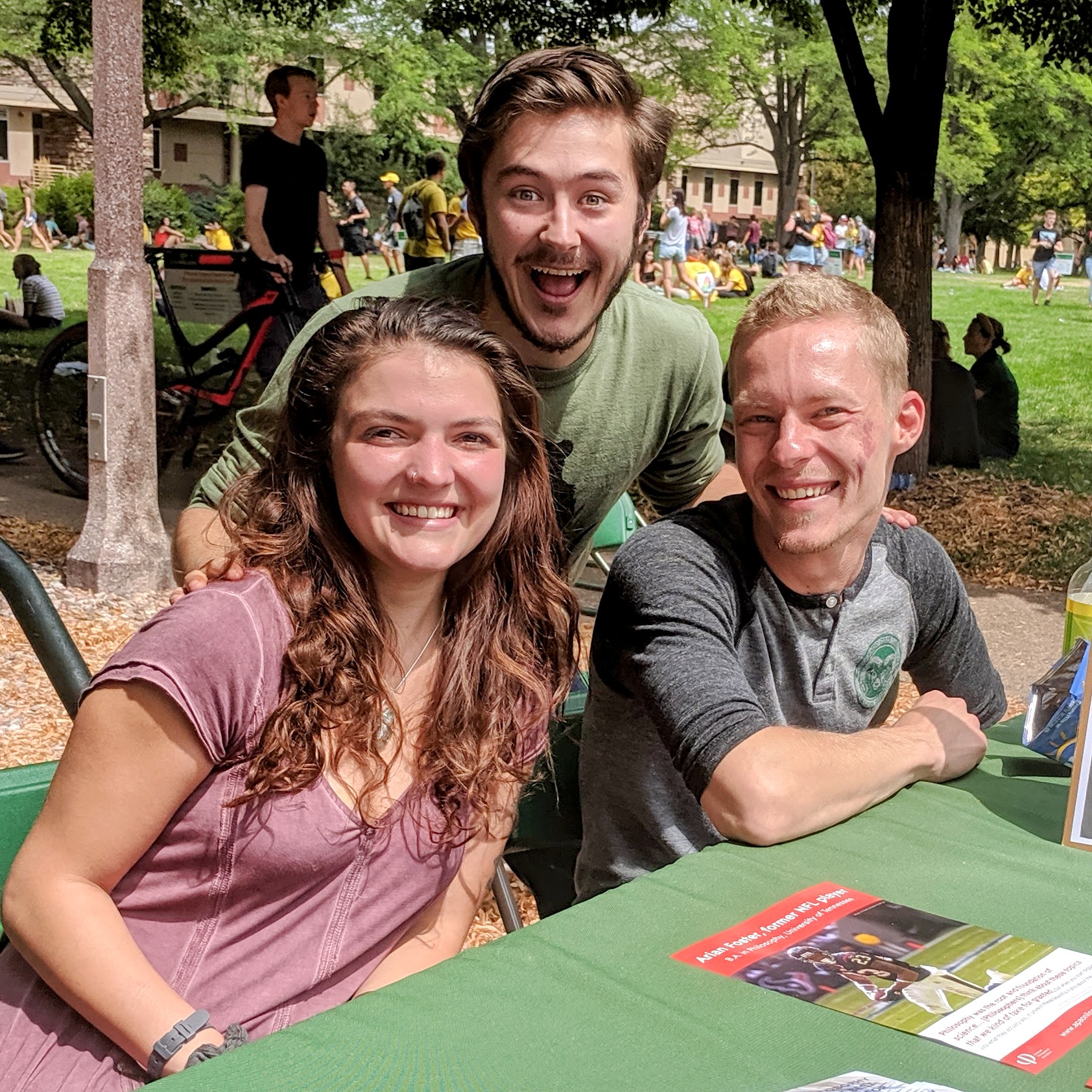 Three people pose together and smile at an outdoor table on a grassy campus quad, with a flyer on the table in front of them.
