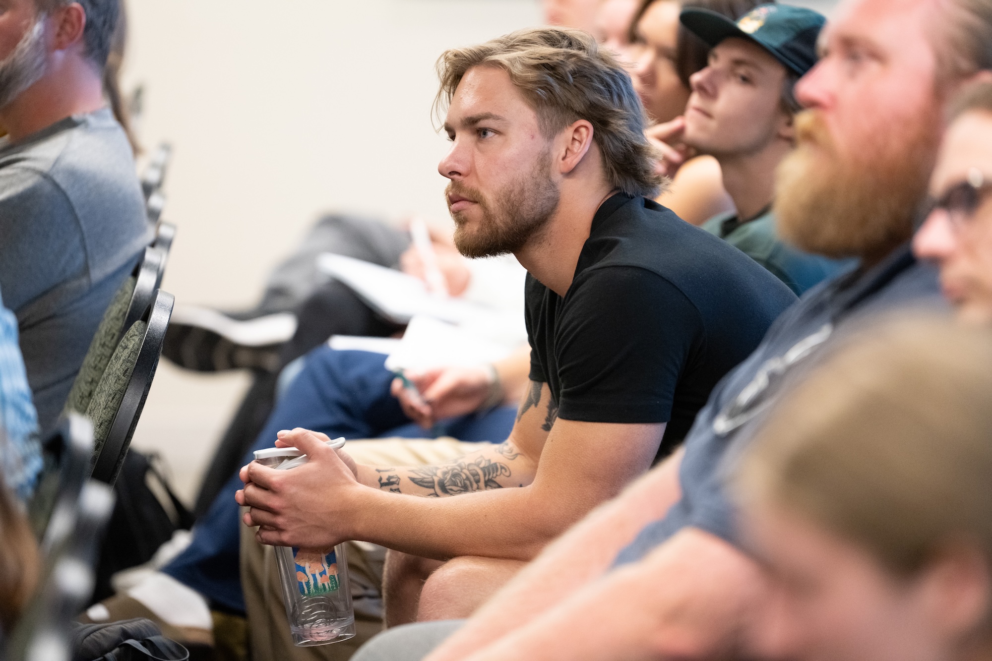A studenet with a beard sits among an audience, holding a water bottle, listening intently with a thoughtful expression.