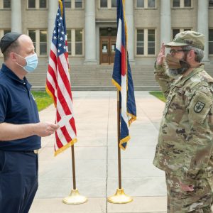 A soldier salutes while receiving an award outdoors, with American flags and a historic building in the background.