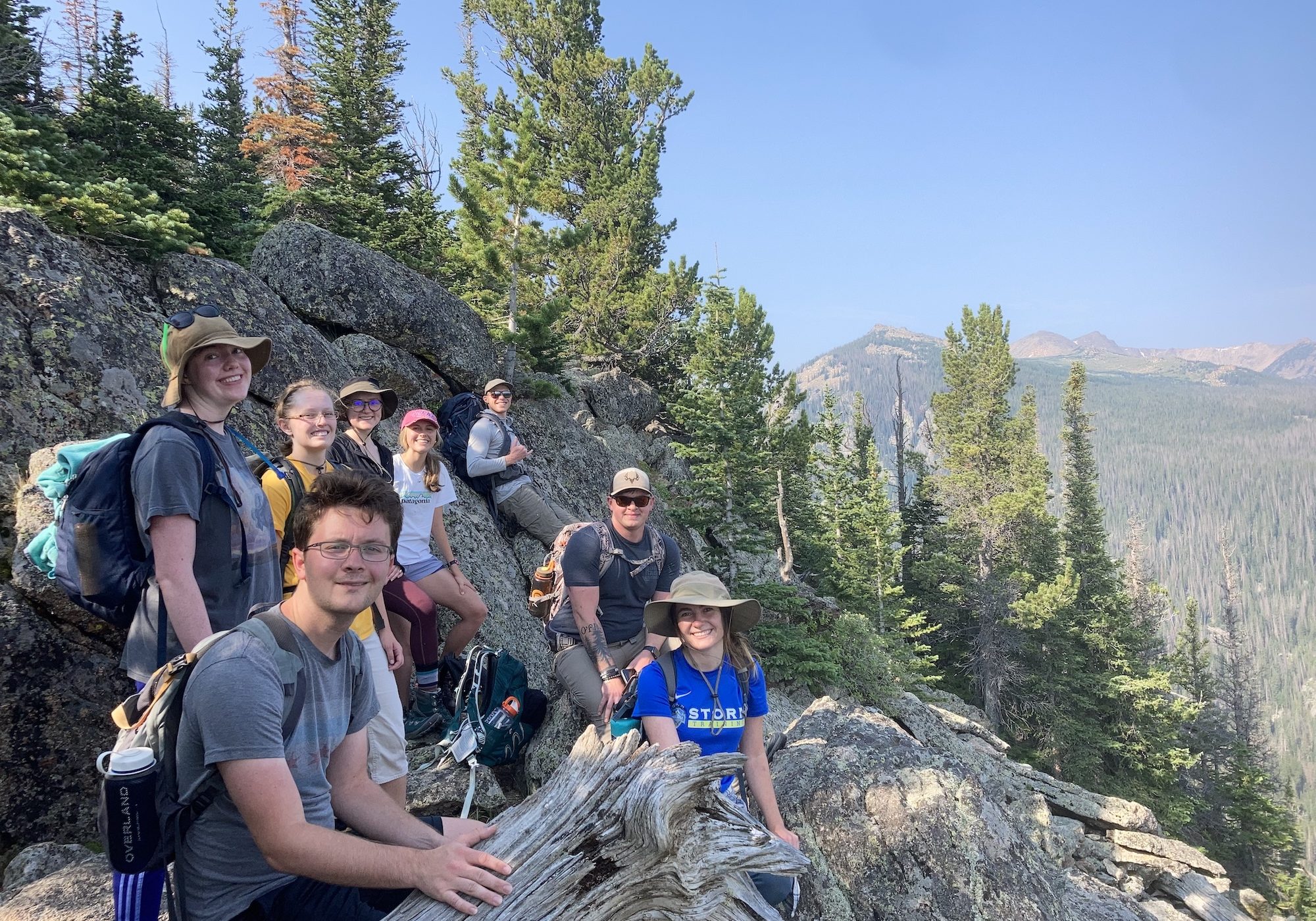 A group of eight people pose together on a rocky mountain outcrop surrounded by pine trees, with a mountain range visible in the distance.
