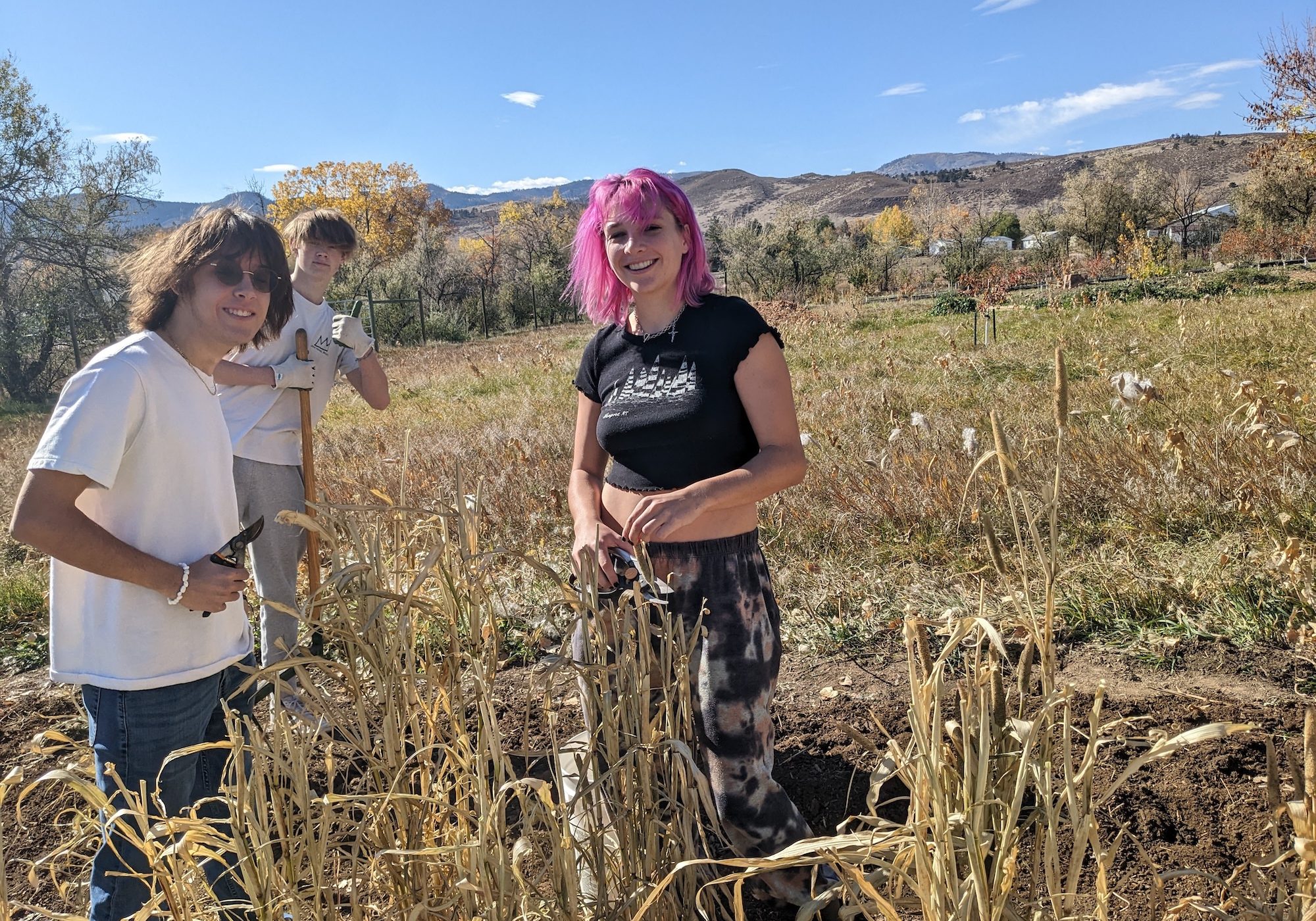 Three students smile while working outdoors in a garden bed, holding pruning shears, with mountains and dry grassland in the background.
